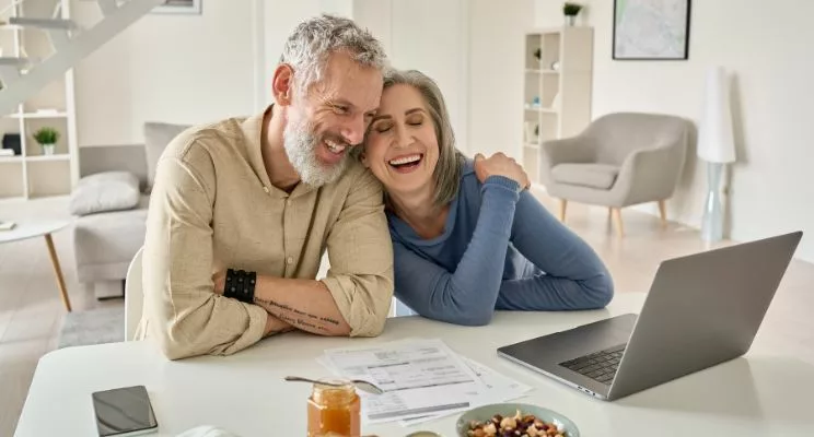 An elderly man and woman are seated in a white room with sleek modern furniture. They are focused on a laptop placed on a table in front of them, exploring and interacting with the digital world.
