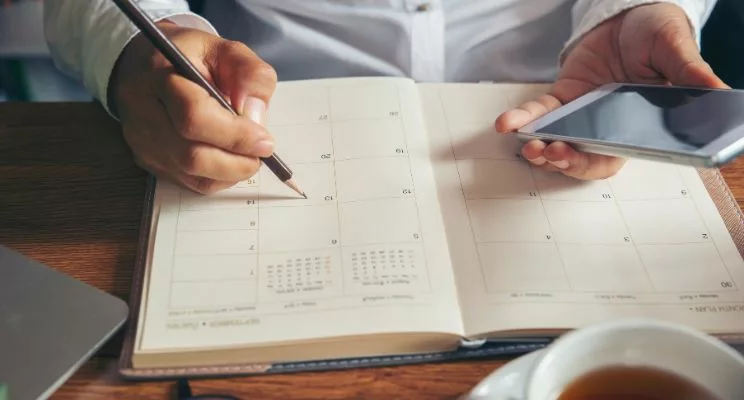 A person is seated at a wooden desk, actively inputting information into a daily planner. They hold a smartphone in their other hand, demonstrating a combination of traditional and digital productivity methods.